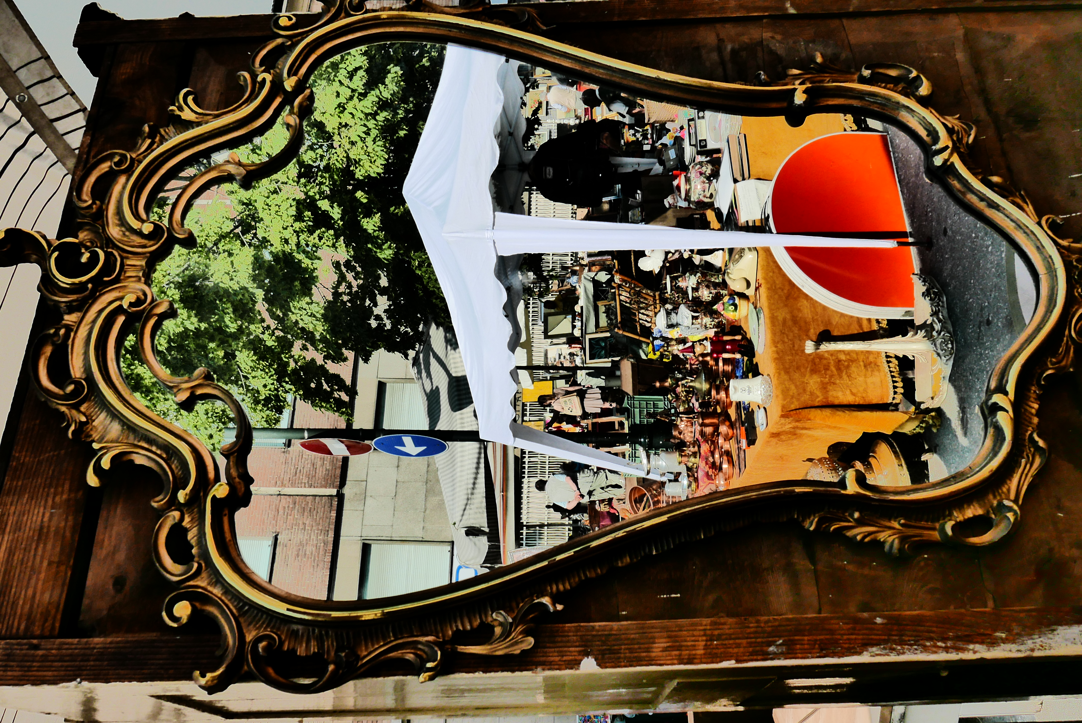 Un brande specchio con la cornice dorata riflette un banchetto di antiquariato sotto un tendone bianco.